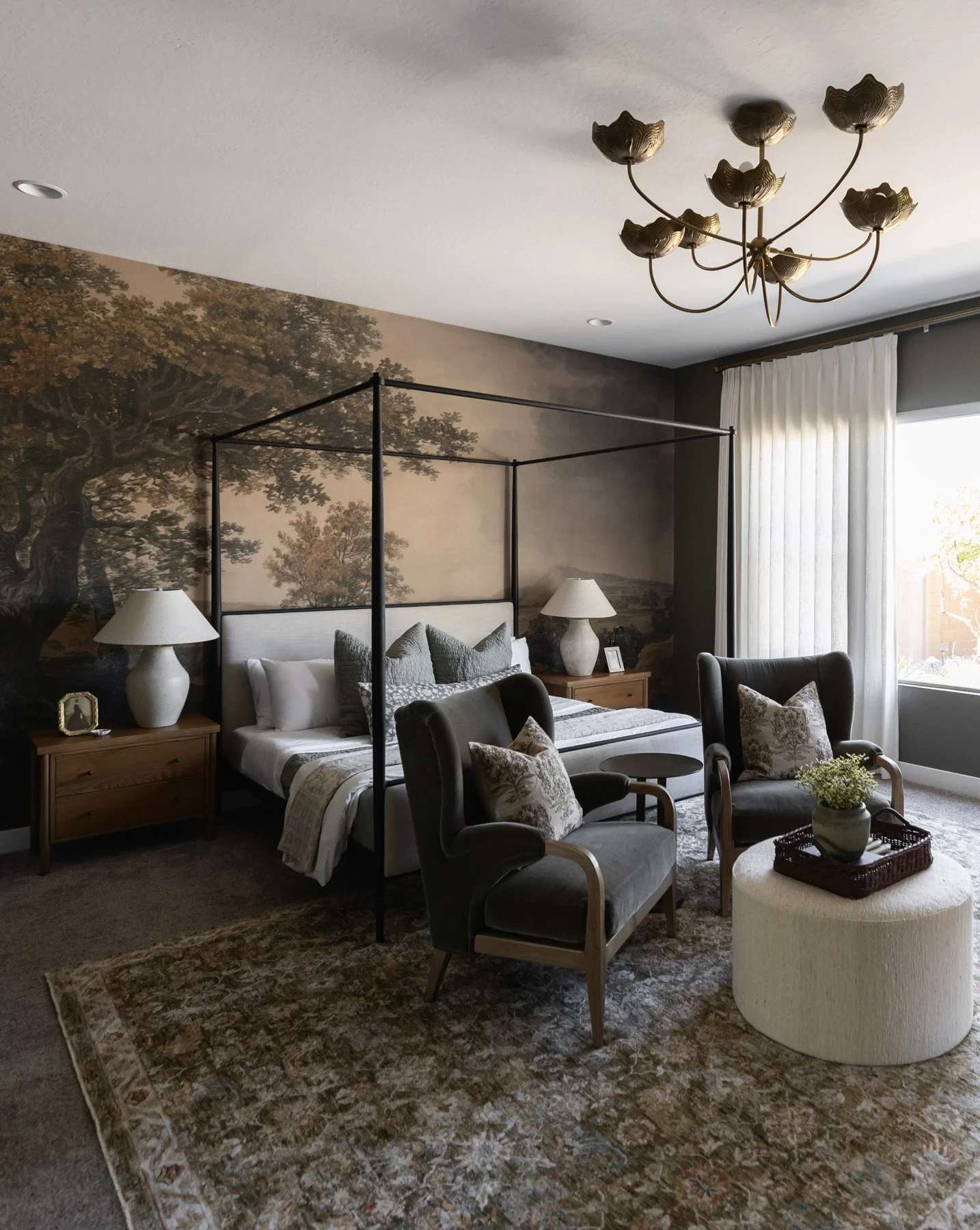 Primary bedroom with black four-poster bed, landscape mural wallpaper, dark olive armchairs, white boucle ottoman, oak bedside tables, white ceramic lamps, vintage-style rug, and brass floral chandelier