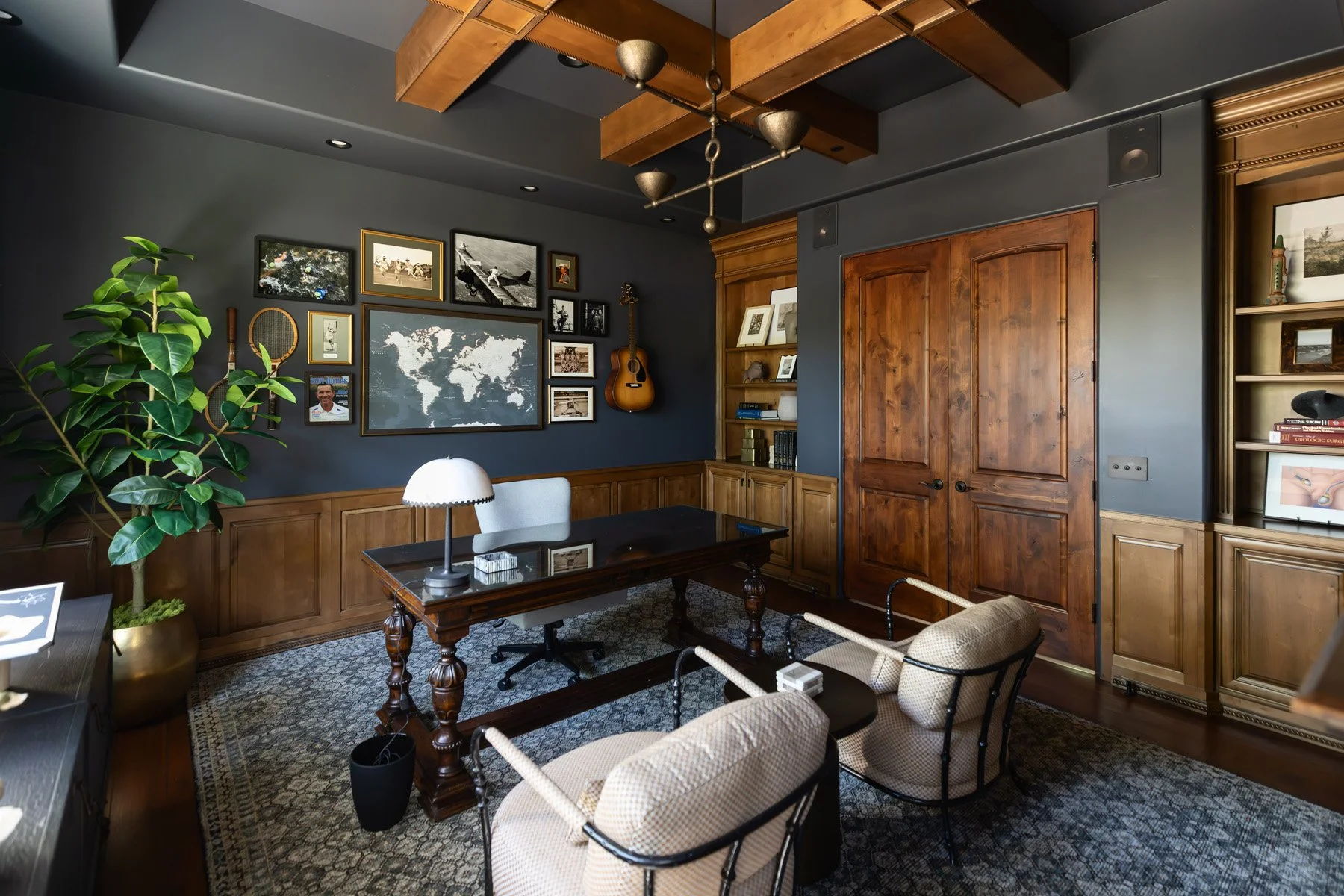 Dark home office with navy walls, dark wood wainscoting, writing desk, white table lamp, potted fiddle leaf fig, framed photo gallery, and upholstered chairs on patterned rug
