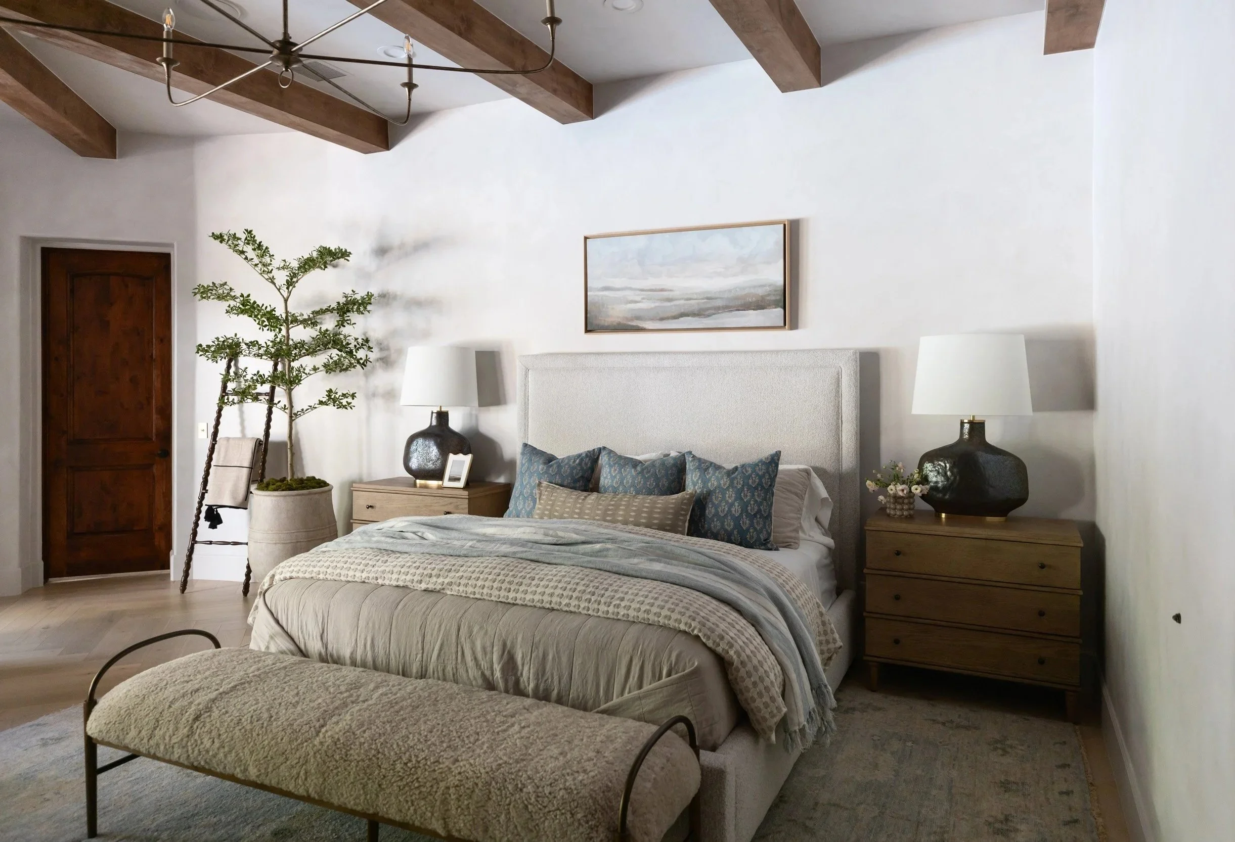 Primary bedroom with upholstered bedhead, blue accent pillows, layered neutral bedding, oak bedside tables, black ceramic lamp, potted tree, and exposed wood ceiling beams