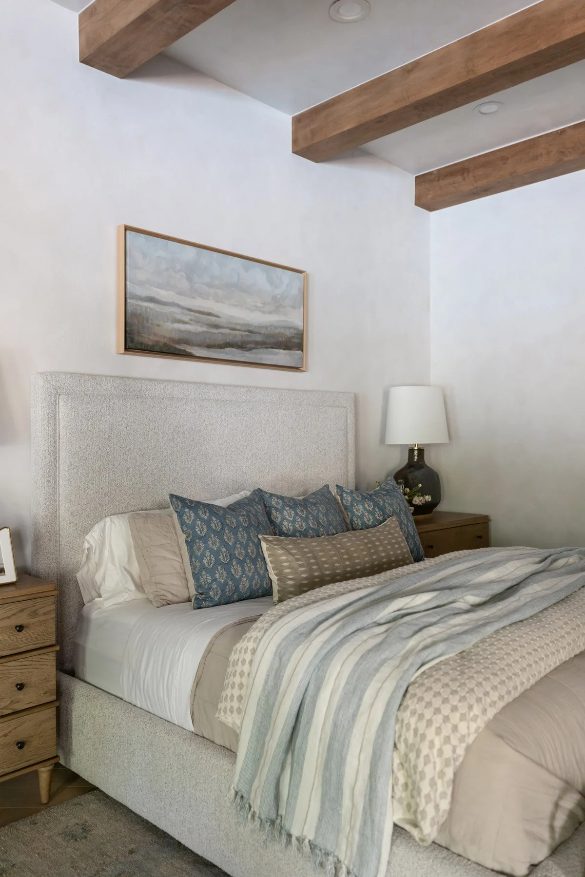 Primary bedroom with upholstered bedhead, blue accent pillows, layered neutral bedding, oak bedside table, white table lamp, landscape artwork, and exposed wood ceiling beams