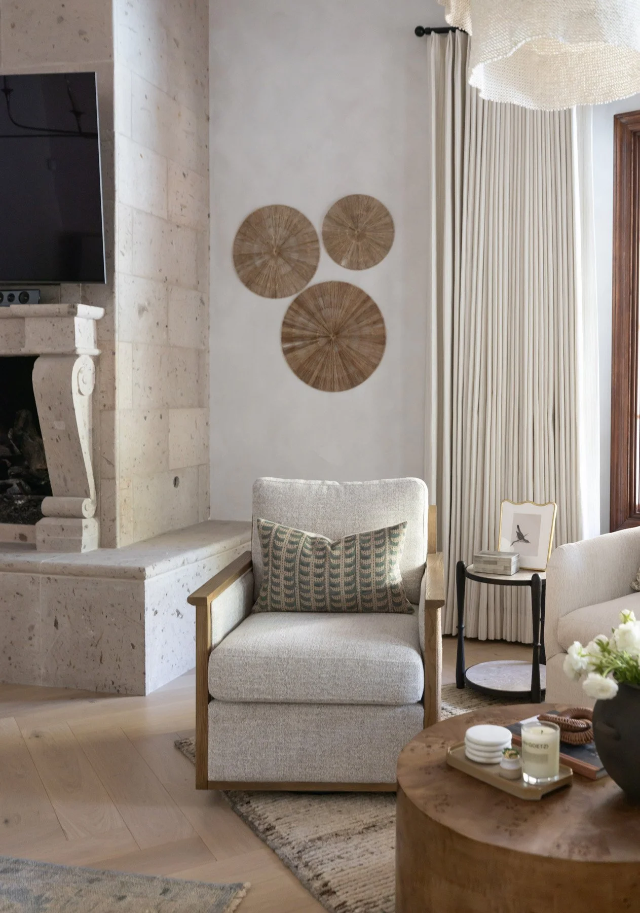 Sitting room with white armchair, black iron side table, circular woven wall art, tall ivory drapery, and wall-mounted TV