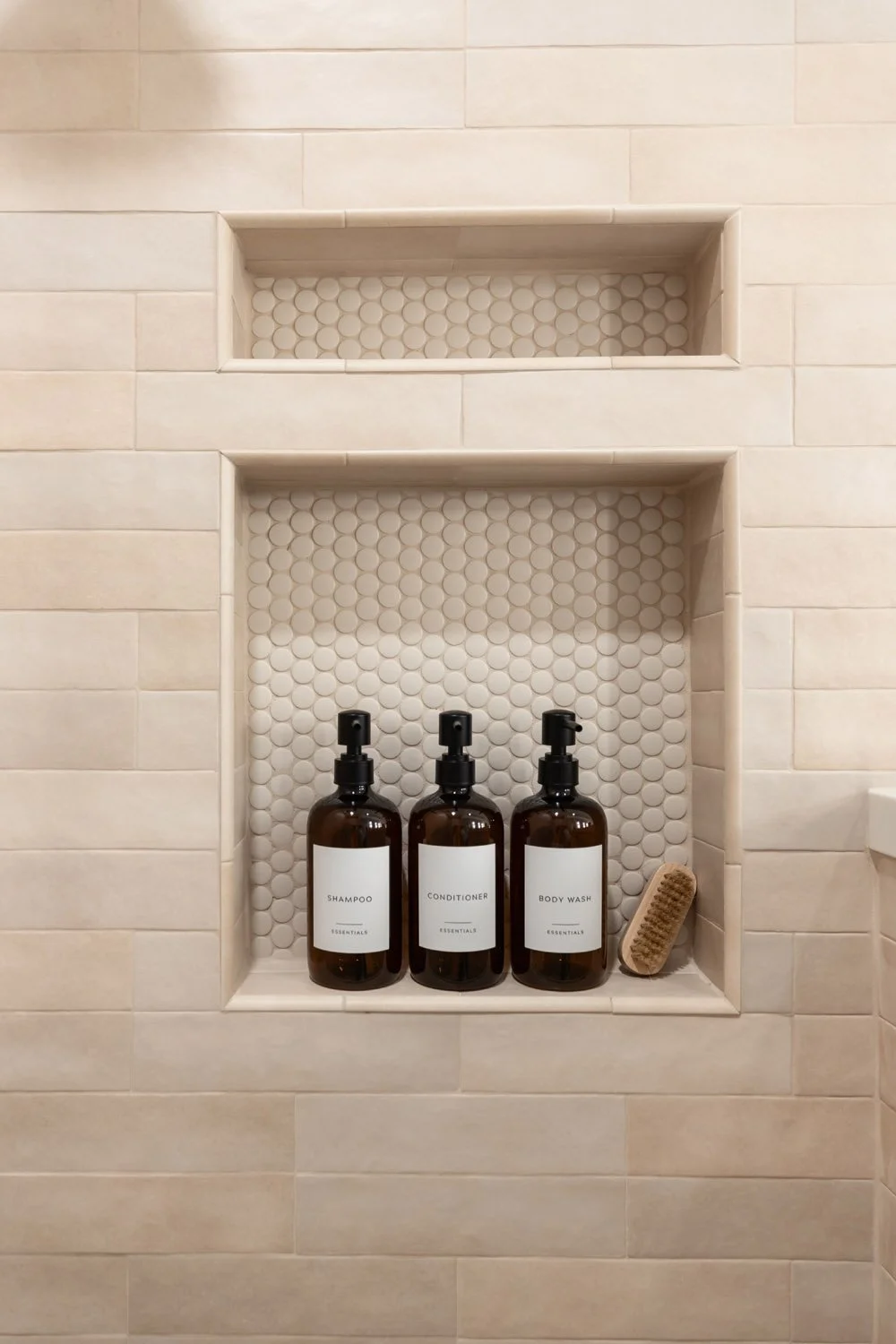 Built-in shower niche with penny round tile backing, styled with amber glass shampoo, conditioner, and body wash bottles and a wooden bath brush
