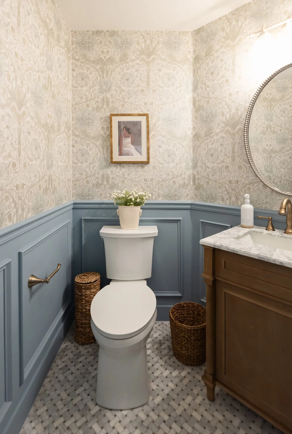 Powder room with blue board and batten wainscoting, botanical wallpaper, dark wood vanity, marble countertop, round mirror, brass fixtures, patterned floor tile, and woven basket