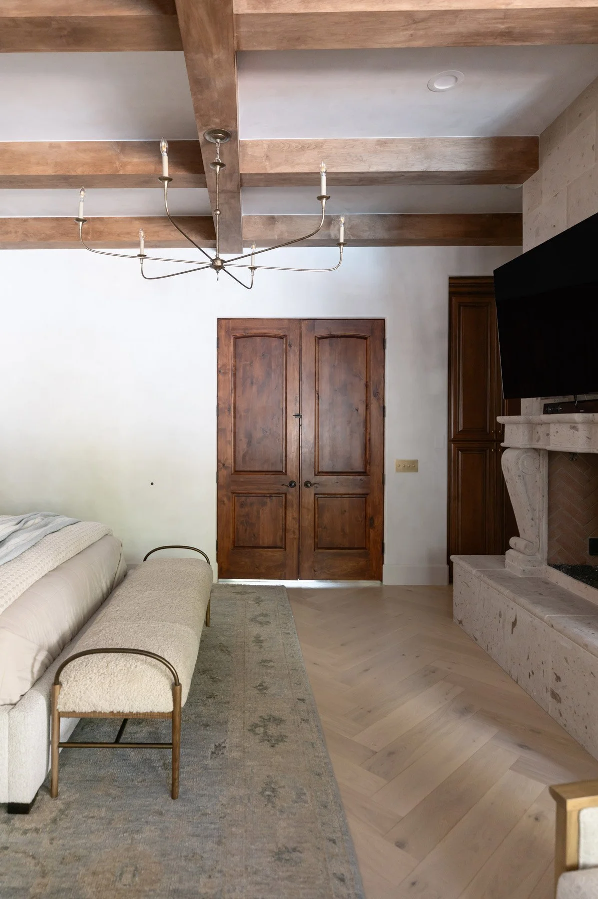 Primary suite with curved bench, white chaise, dark wood double doors, wall-mounted TV, and exposed wood ceiling beams