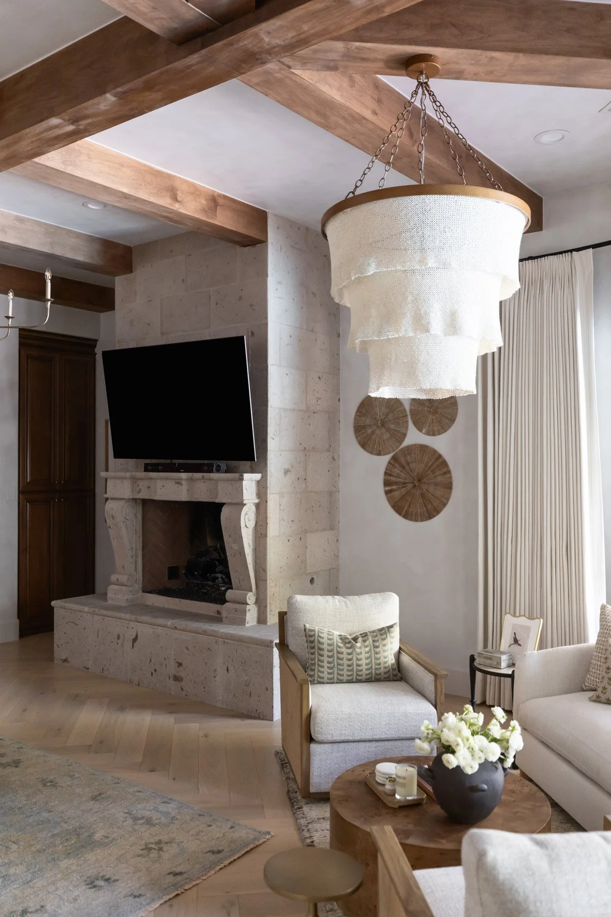 Primary suite with white armchairs, woven fringe pendant light, stone fireplace, circular woven wall art, wall-mounted TV, and neutral drapery