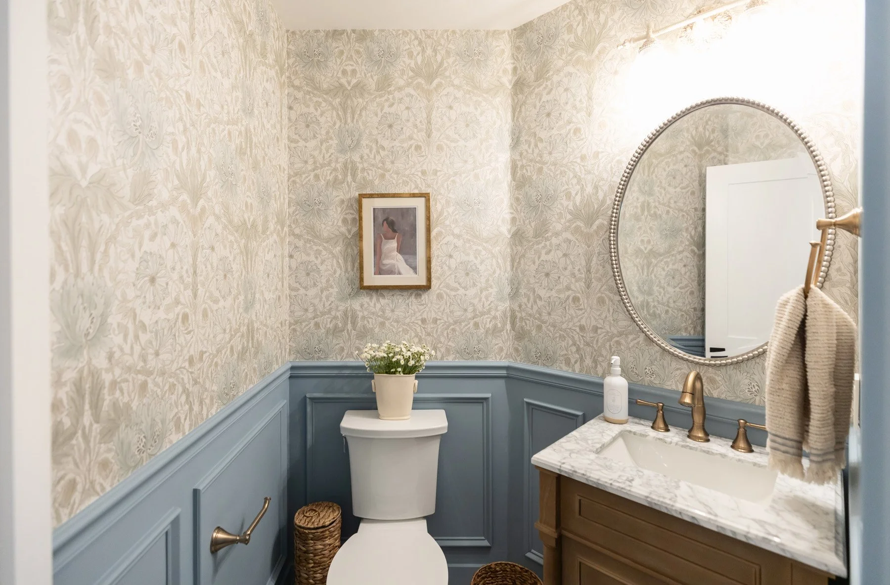 Powder room with blue board and batten wainscoting, botanical wallpaper, dark wood vanity, marble countertop, oval mirror, brass fixtures, and framed artwork