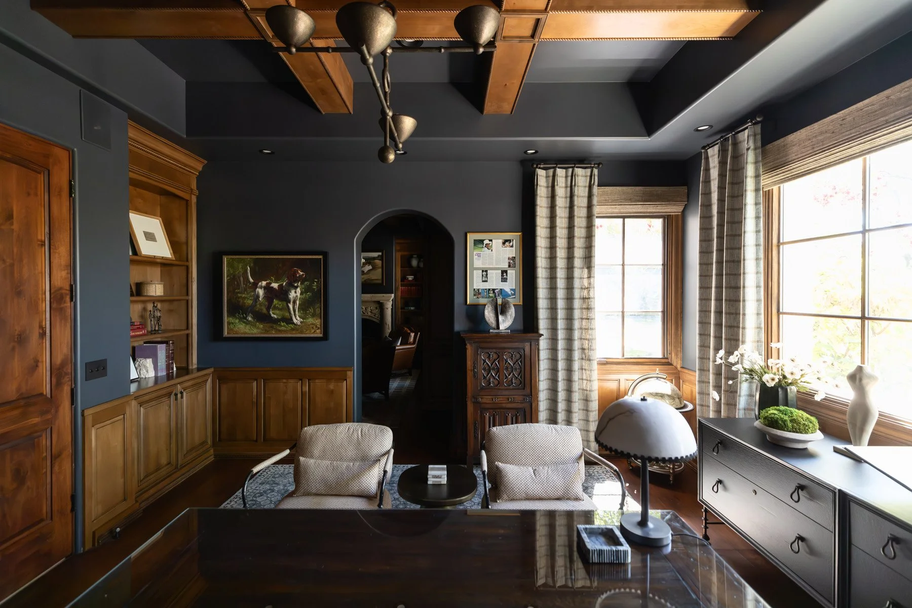 Dark home office with navy walls, dark wood wainscoting, writing desk, white table lamp, arched built-in cabinetry, large windows with check drapery, and brass ceiling light
