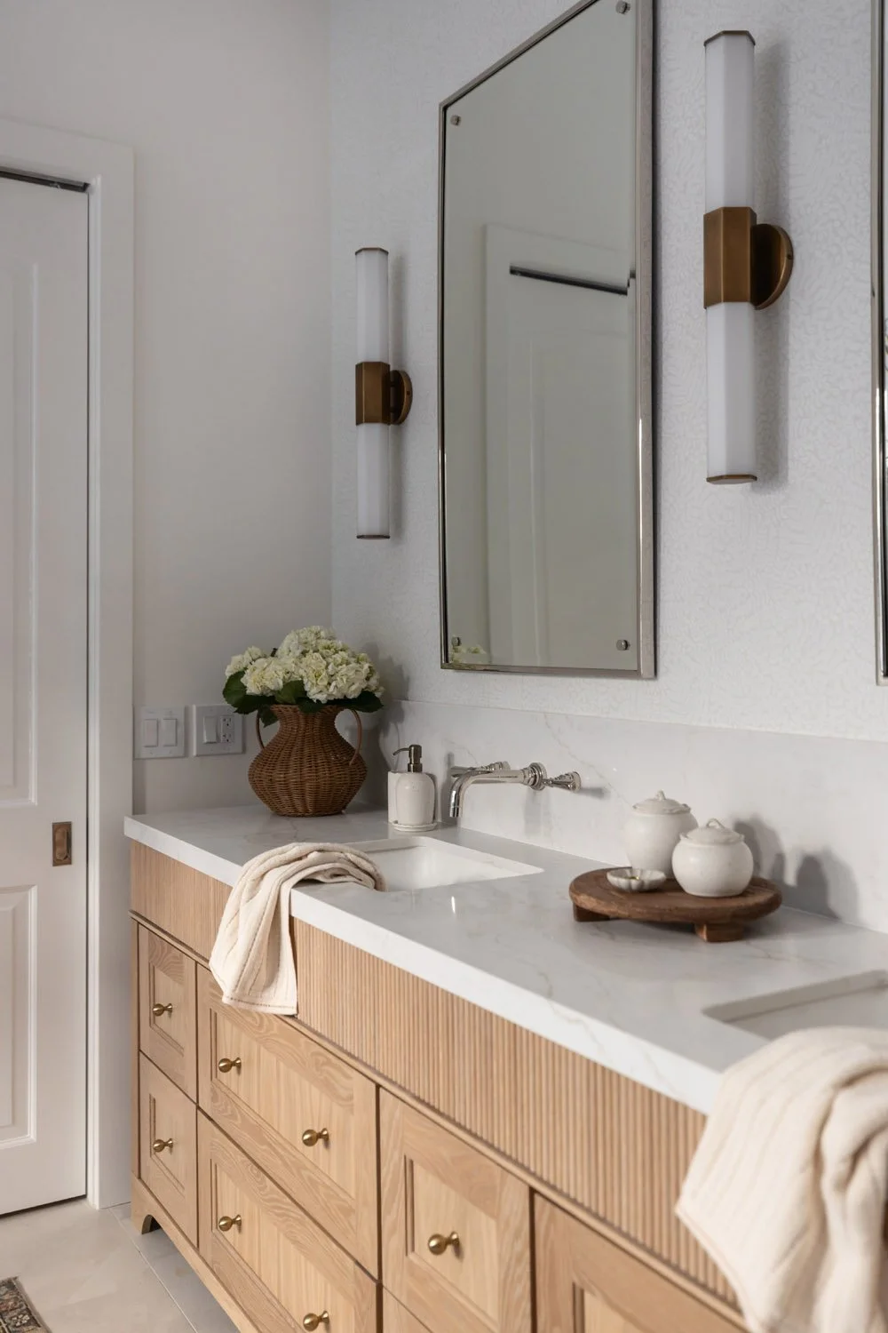 Light oak vanity with white quartz countertop, brass and nickel fixtures, fresh floral arrangement, and brass and white glass wall sconces flanking a black-framed mirror