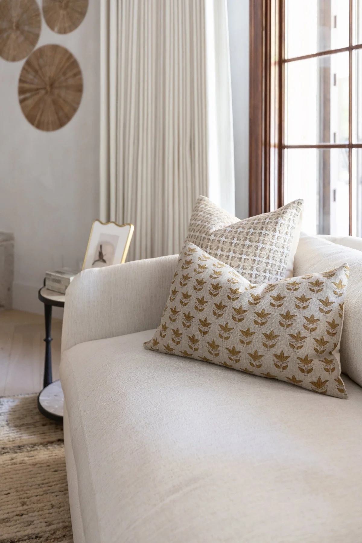 White chaise lounge with block print accent pillow, circular woven wall art, and dark wood French windows with neutral drapery