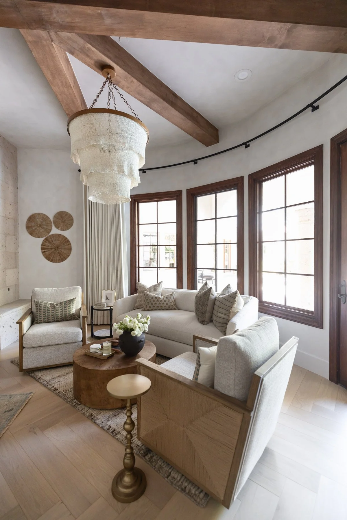 Sitting room with white armchair, brass side table, circular woven wall art, woven fringe pendant, dark wood French windows, and exposed wood ceiling beams