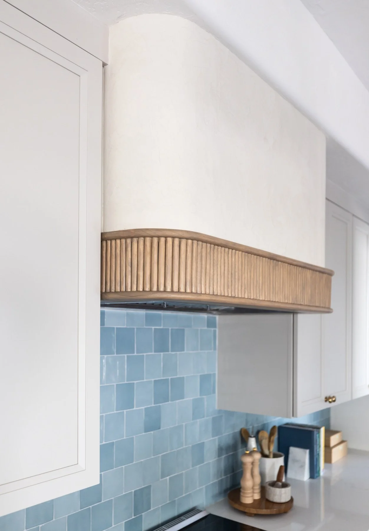 Close-up of woven fluted range hood above blue zellige tile backsplash with white shaker cabinetry and brass hardware