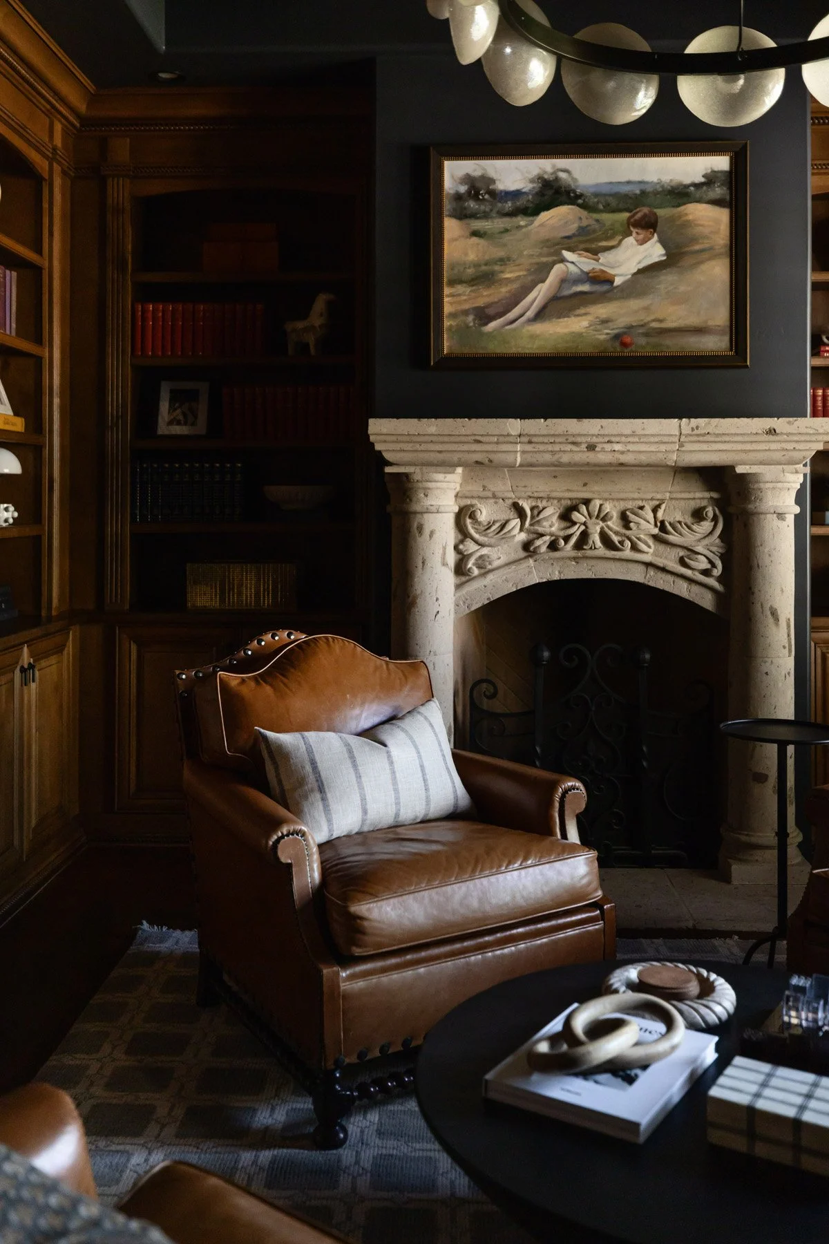 Ornate stone fireplace with framed landscape artwork above, leather armchair, coffee table with decorative objects, and dark wood built-in bookshelves