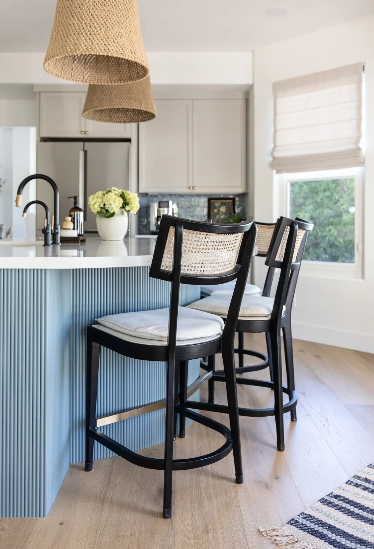 Kitchen with blue fluted island, black cane bar stools, woven pendant light, white shaker cabinetry, blue zellige tile backsplash, and woven roman blinds