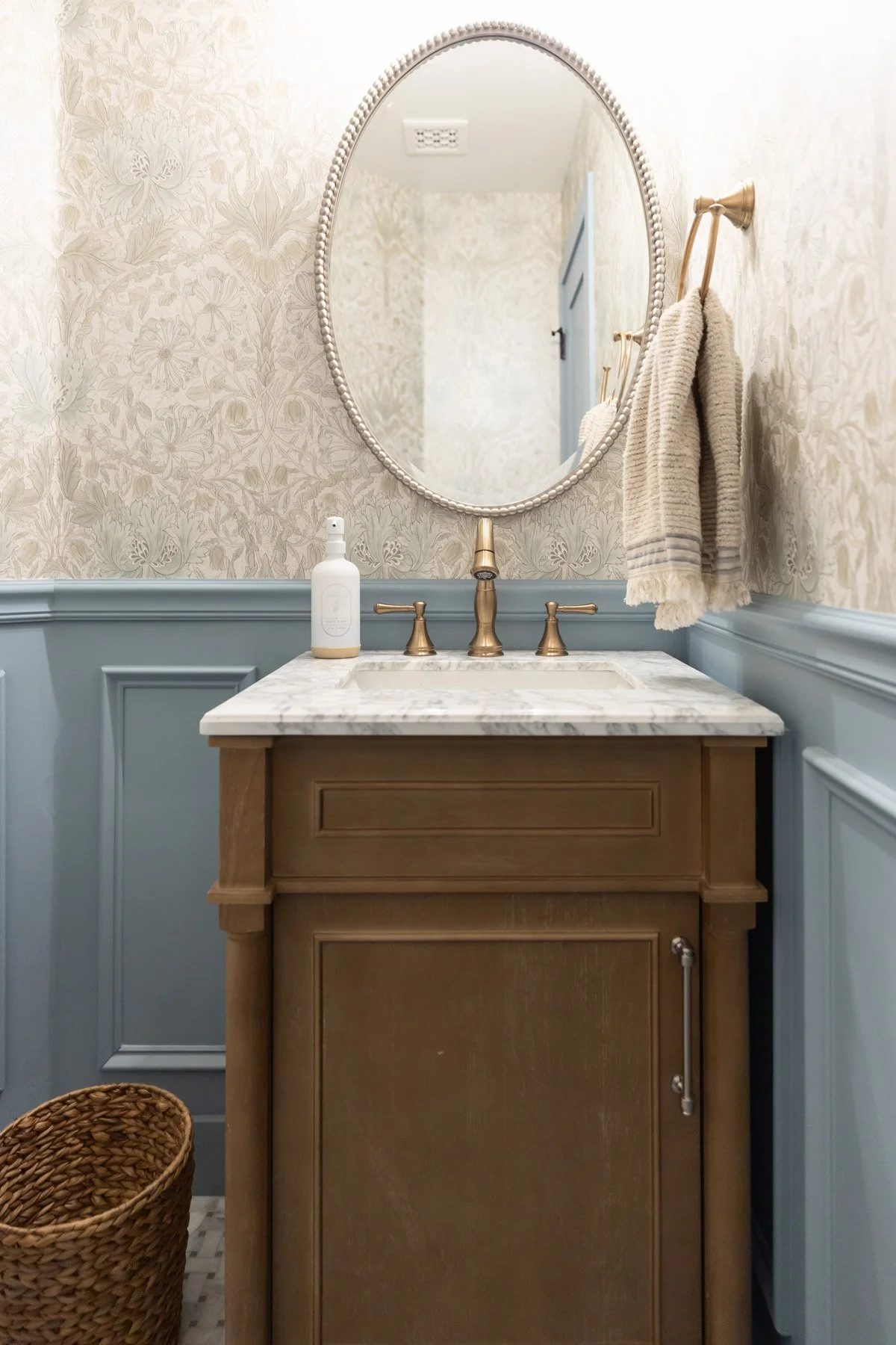 Coastal powder room vanity with dark wood cabinetry, marble countertop, oval mirror, brass fixtures, botanical wallpaper, and blue board and batten wainscoting
