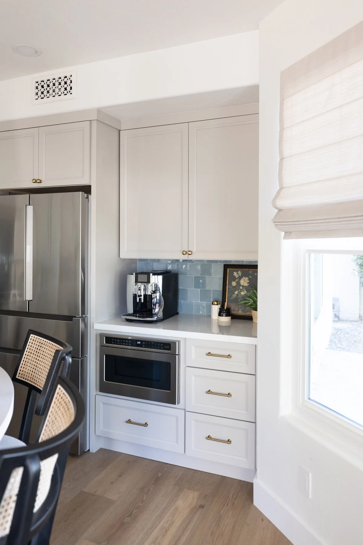 Kitchen appliance wall with white shaker cabinetry, brass hardware, blue zellige tile backsplash, built-in microwave, stainless steel fridge, and cane bar stool