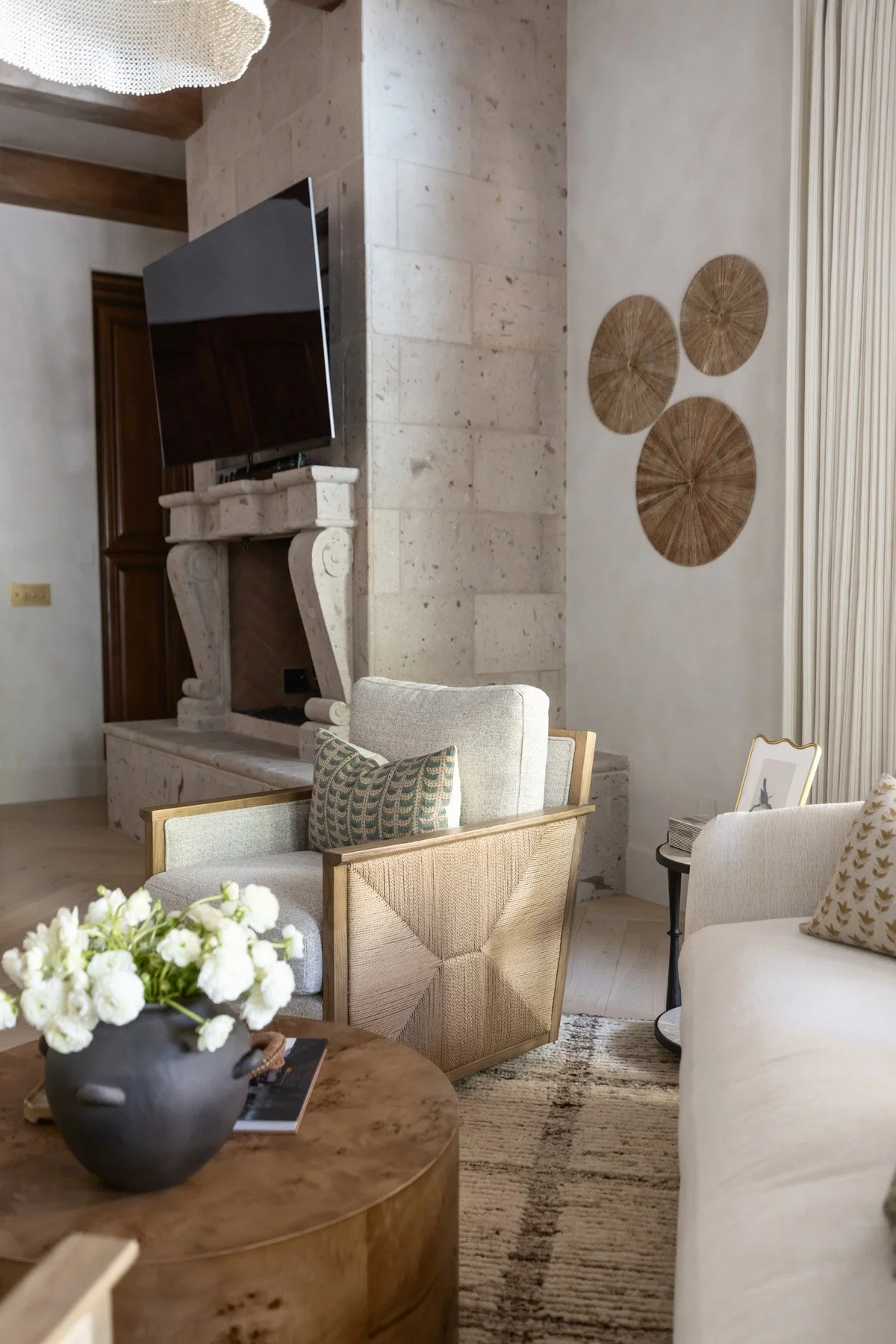 Primary suite sitting area with wall-mounted TV, circular woven wall art, white armchair, round wood slab coffee table, black ceramic vase, and stone fireplace surround