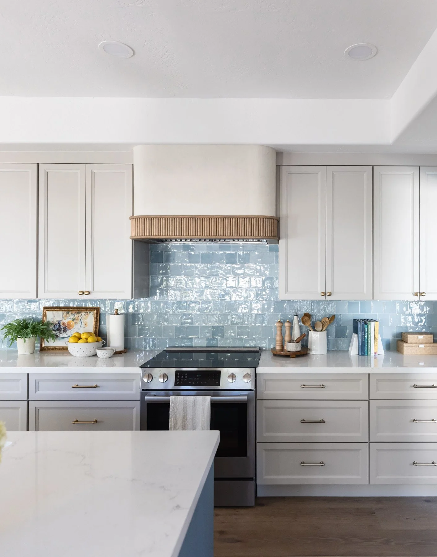 Kitchen with white shaker cabinetry, blue zellige tile backsplash, gold hardware, woven range hood, blue fluted island, and stainless steel range