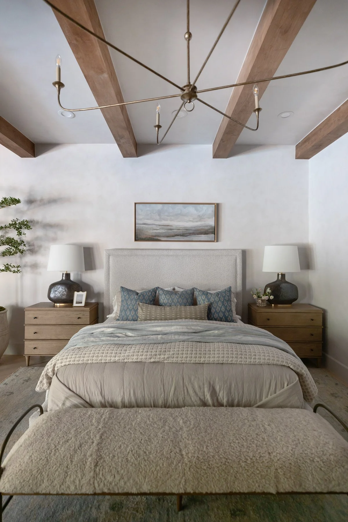 Primary bedroom with upholstered bedhead, layered neutral bedding, oak bedside table, black ceramic lamp, and sculptural black branch chandelier with exposed wood ceiling beams