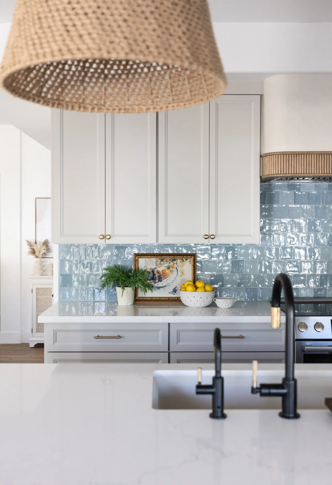 Kitchen island detail with white quartz countertop, black faucet, blue zellige tile backsplash, white shaker cabinetry, woven pendant light, and framed artwork on counter
