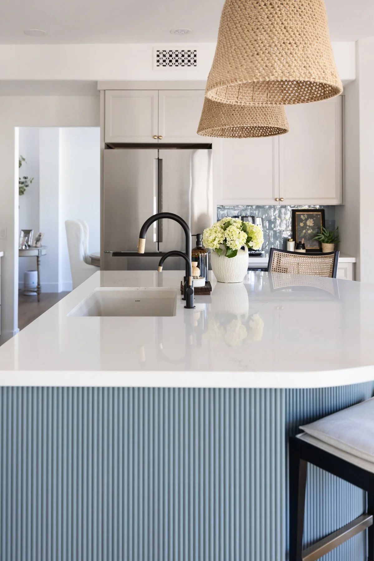 Coastal kitchen island detail with white quartz countertop, black faucet, white floral arrangement, woven pendant light, and blue fluted island base