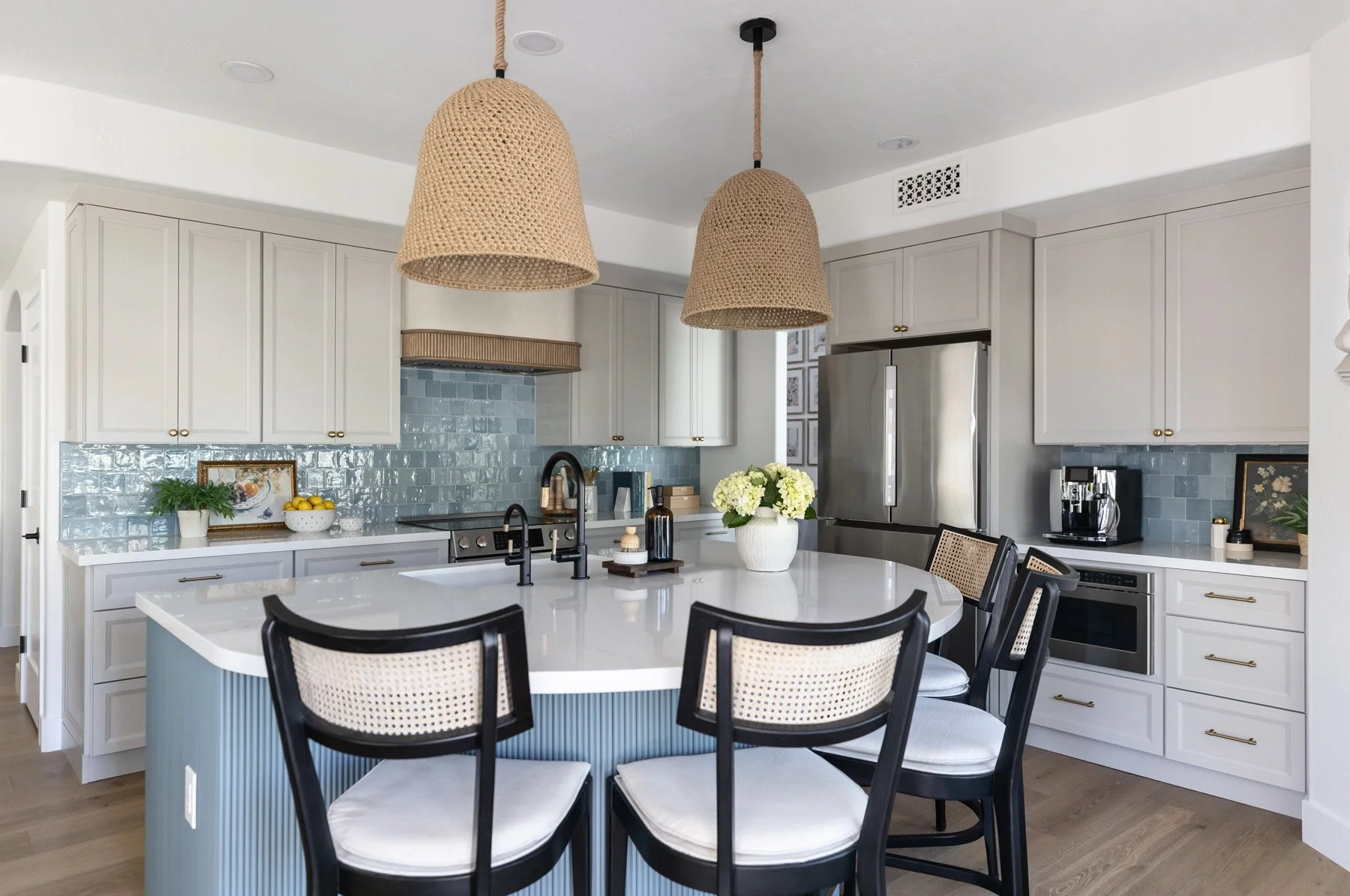 Coastal kitchen overview with blue fluted island, black cane bar stools, woven pendant lights, white shaker cabinetry, blue zellige tile backsplash, and stainless steel fridge