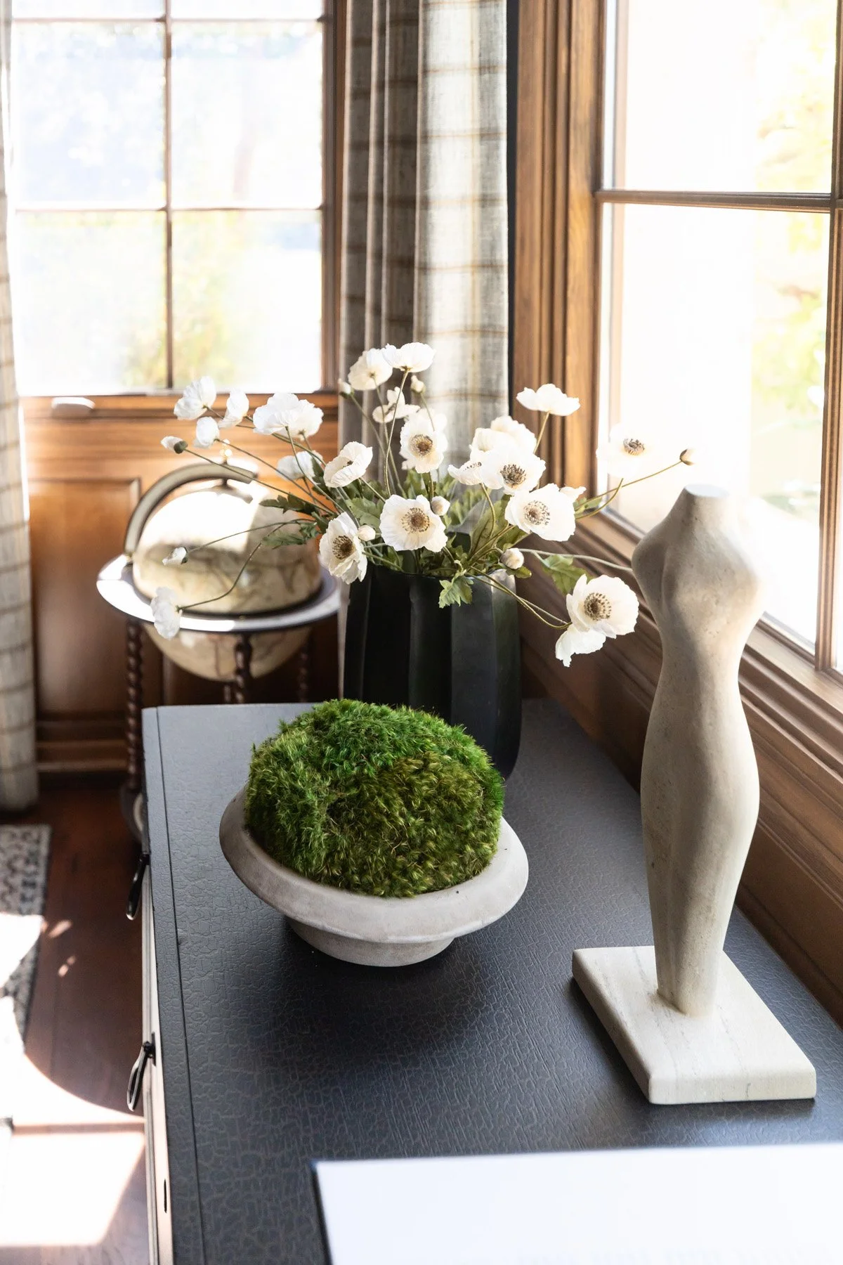 Dark coffee table styled with white floral arrangement, moss bowl, and sculptural white object beside window with check drapery