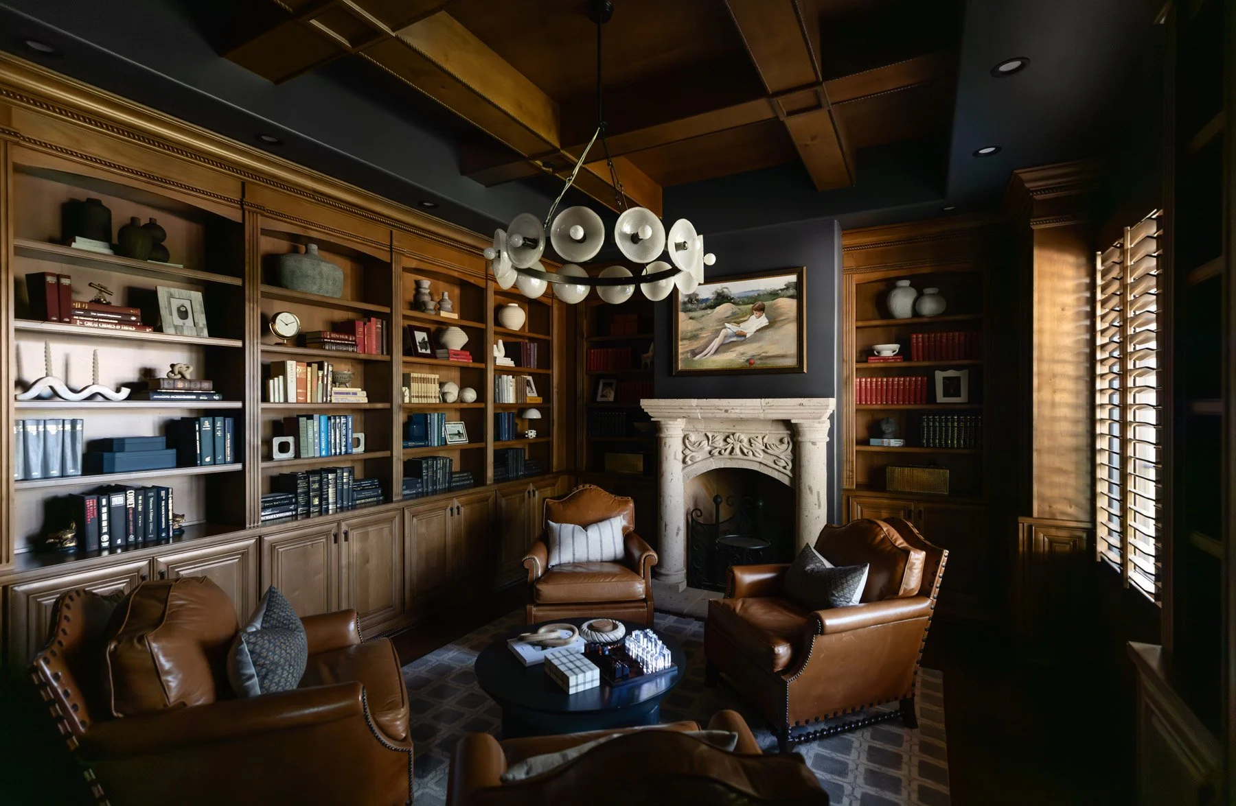 Dark study with leather armchairs, ornate stone fireplace, built-in dark wood bookshelves, and woven roman blinds