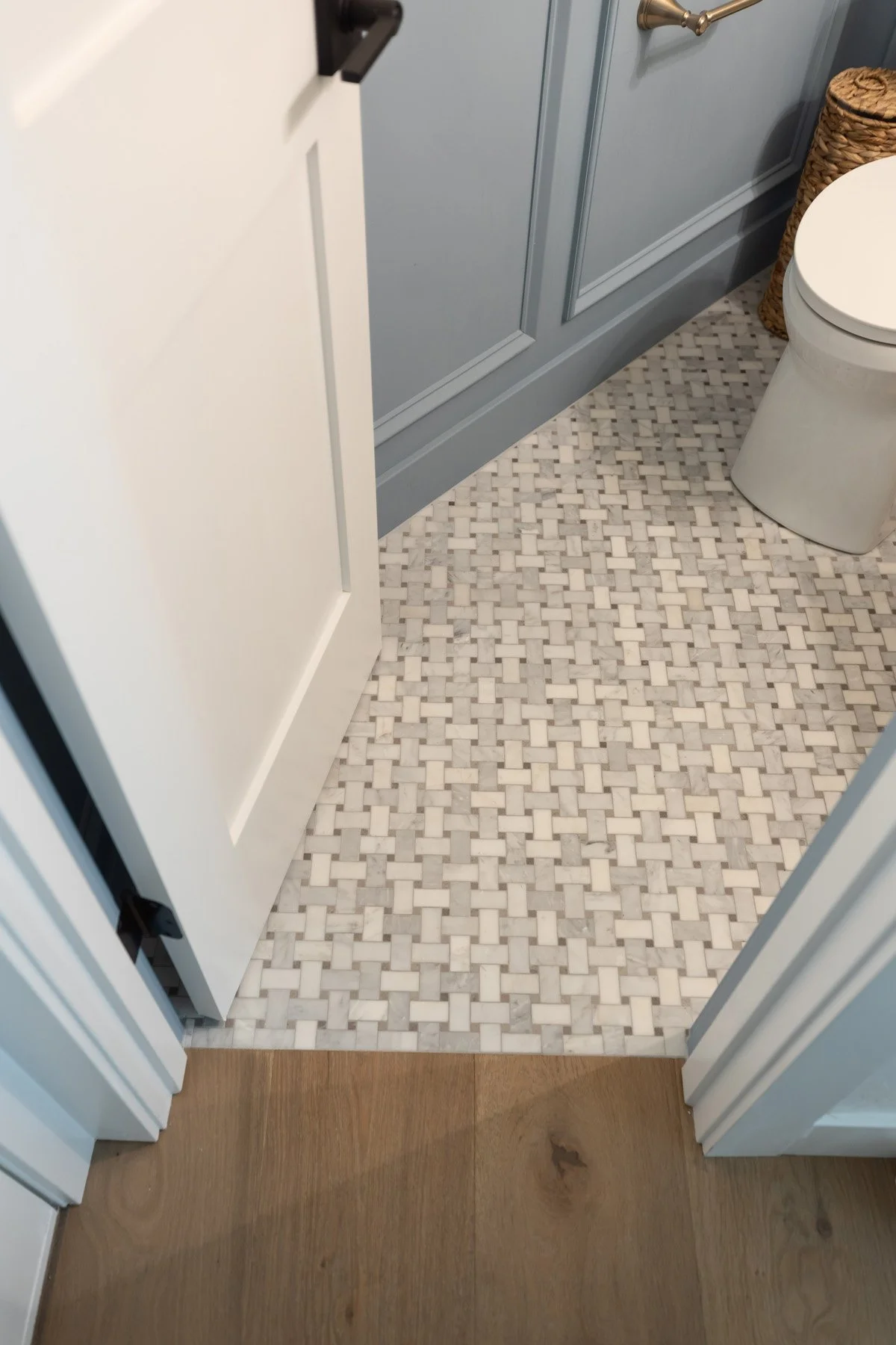 Coastal powder room floor transition detail with patterned mosaic tile, blue painted skirting, and light oak timber floor beyond door threshold