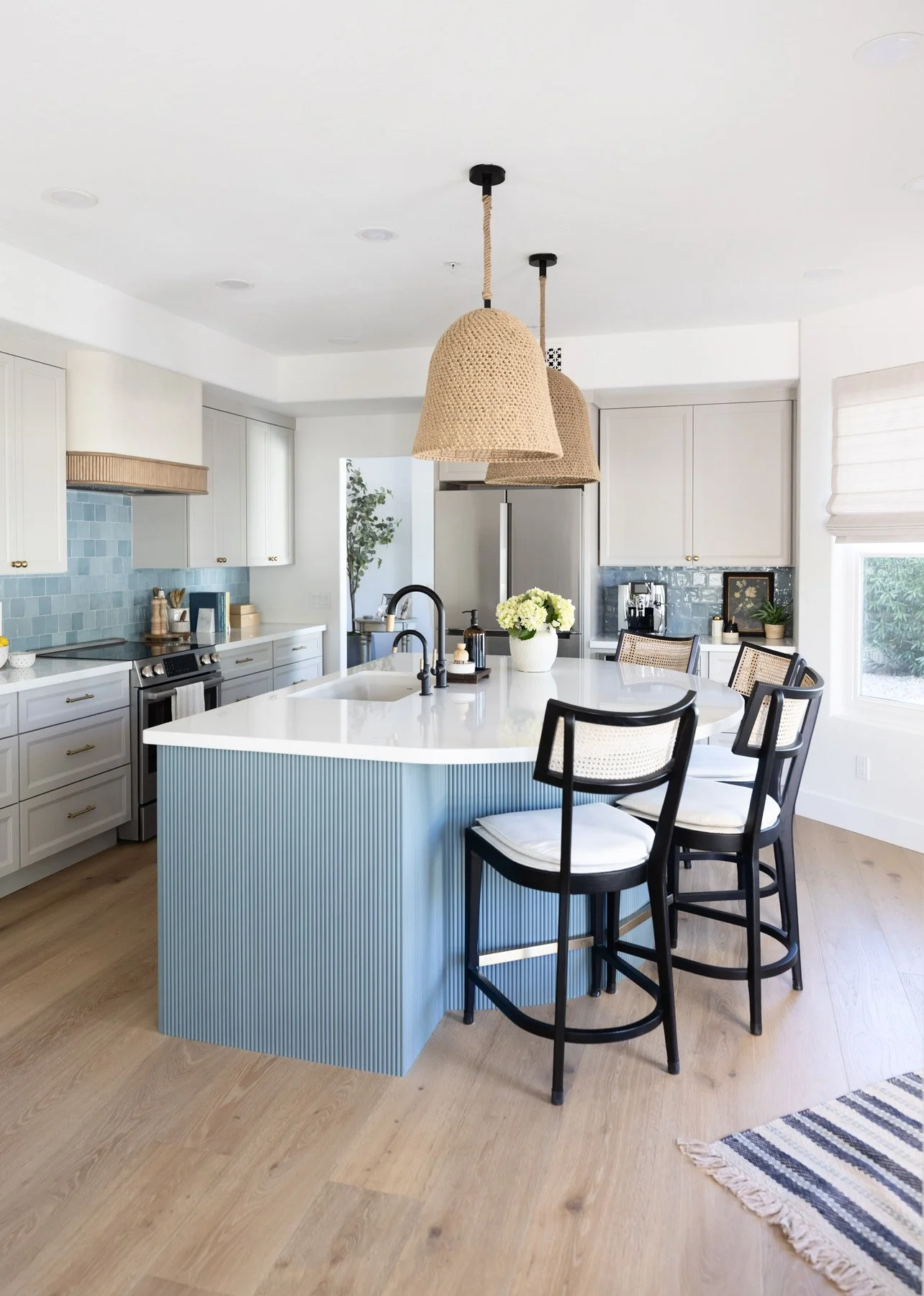 Coastal kitchen with blue fluted island, white quartz countertop, black cane bar stools, woven pendant light, white shaker cabinetry, and striped runner rug on light oak floors