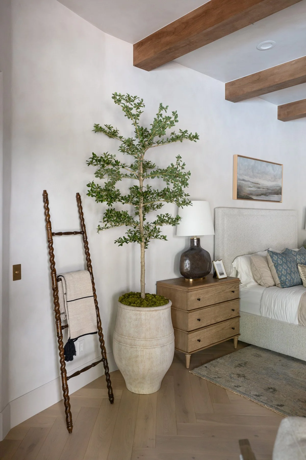 Bedroom corner with lean ladder shelf, large potted tree, oak dresser, black ceramic lamp, and landscape artwork on white wall