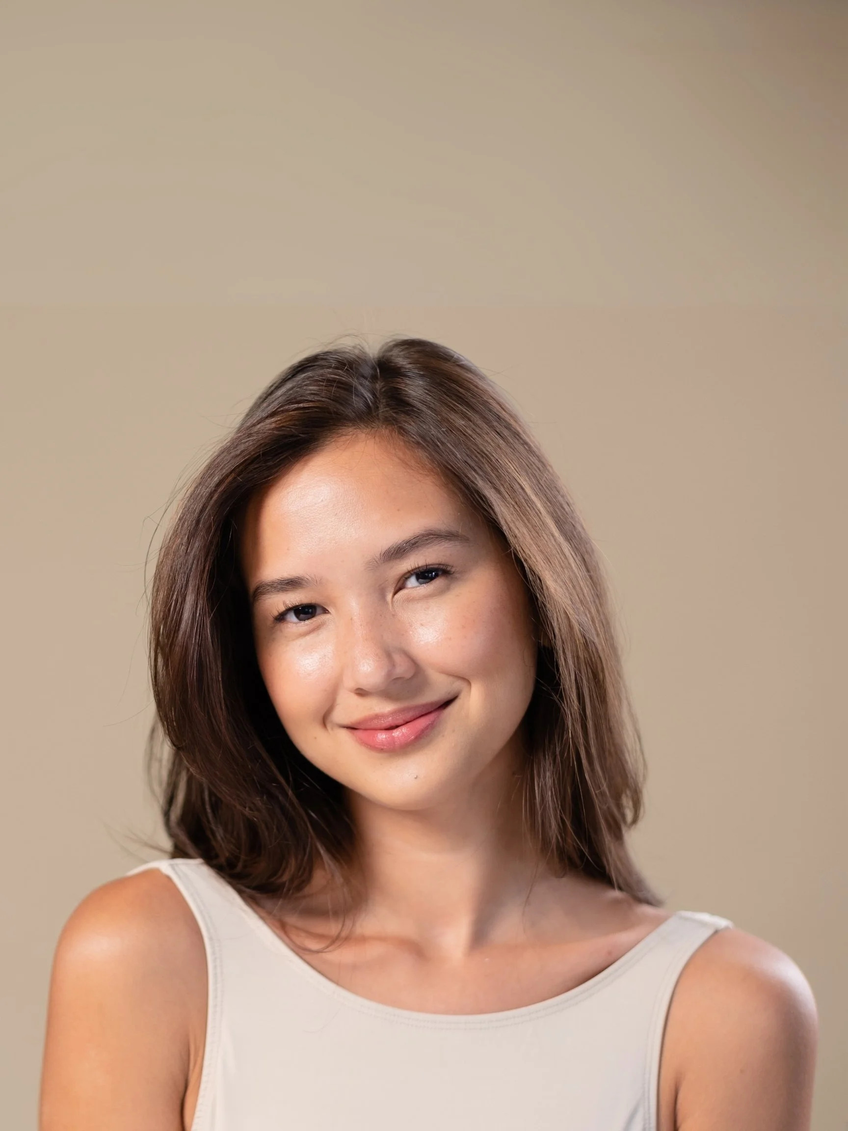 A young woman with shoulder-length brown hair smiling at the camera, wearing a sleeveless beige top against a plain beige background.