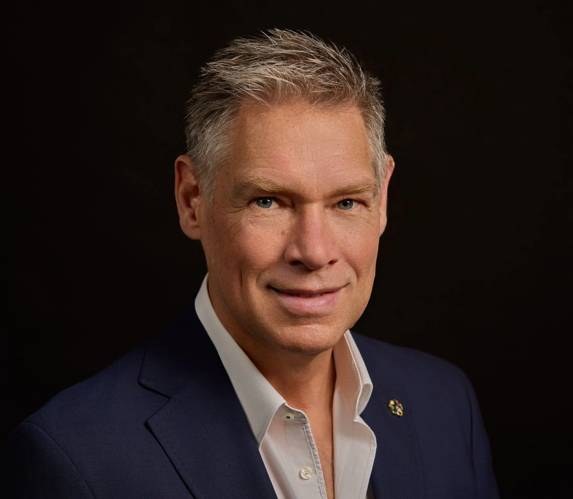 A professional headshot of a middle-aged man with short, blonde hair, wearing a dark suit and white shirt, against a black background.