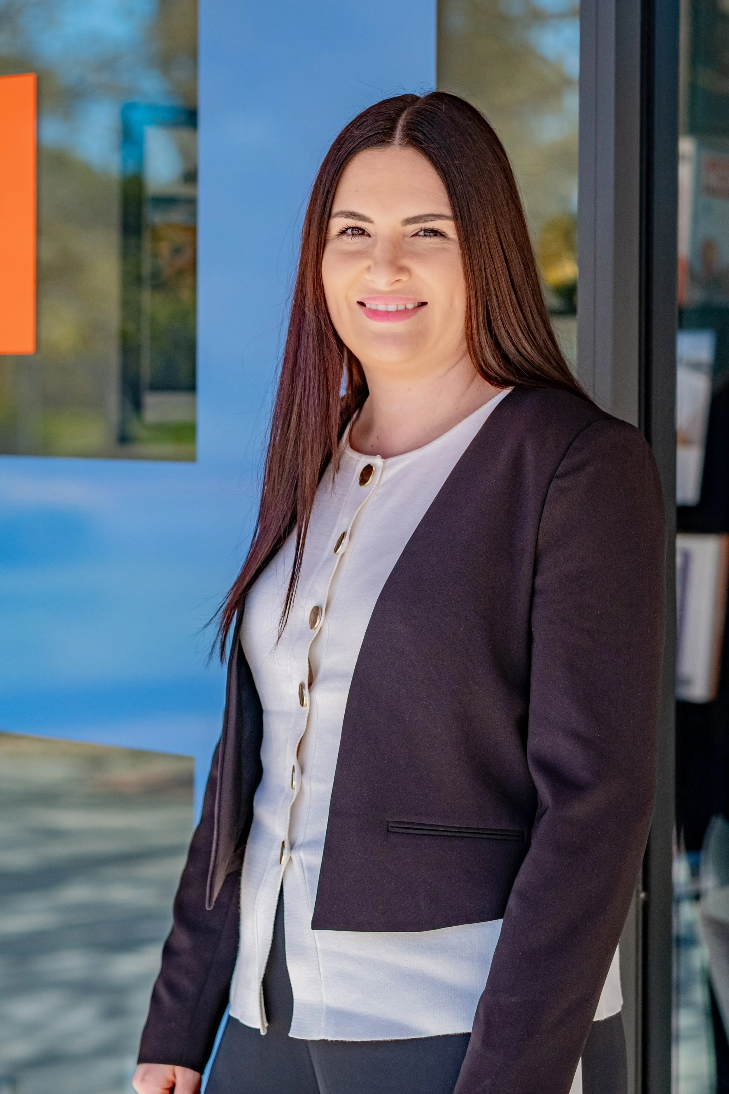 A woman with long brown hair smiling and winking, dressed in a white blouse and black blazer, standing outside near a blue building.