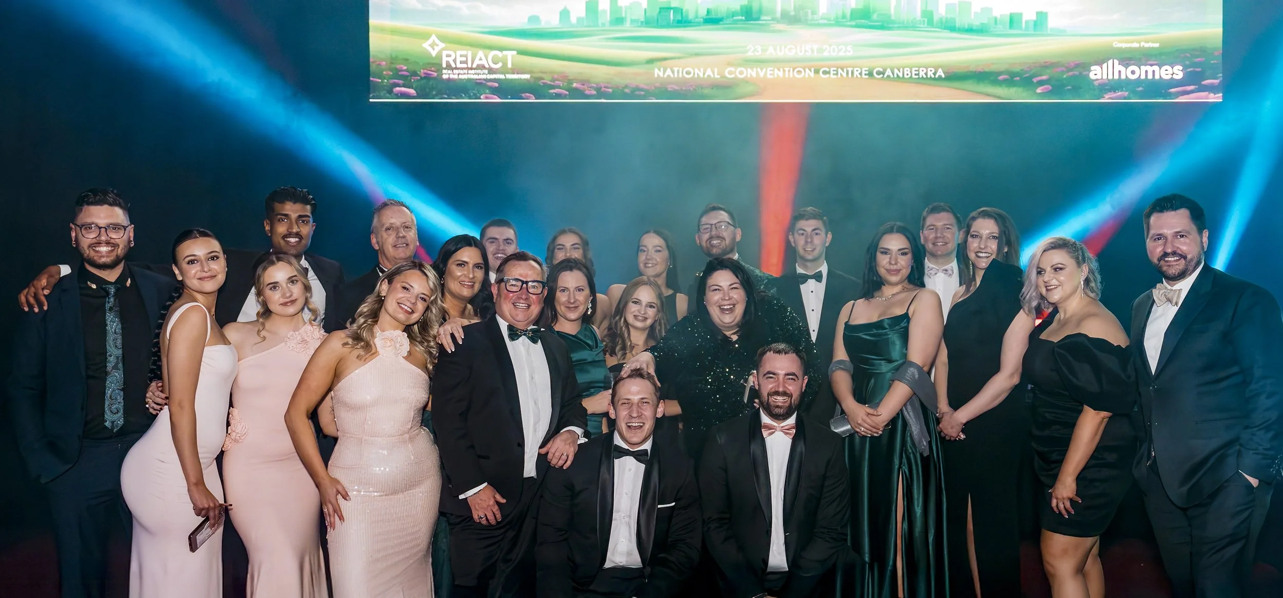 Group of well-dressed men and women smiling at a formal event on a stage, with a banner overhead displaying the date, location, and event name.