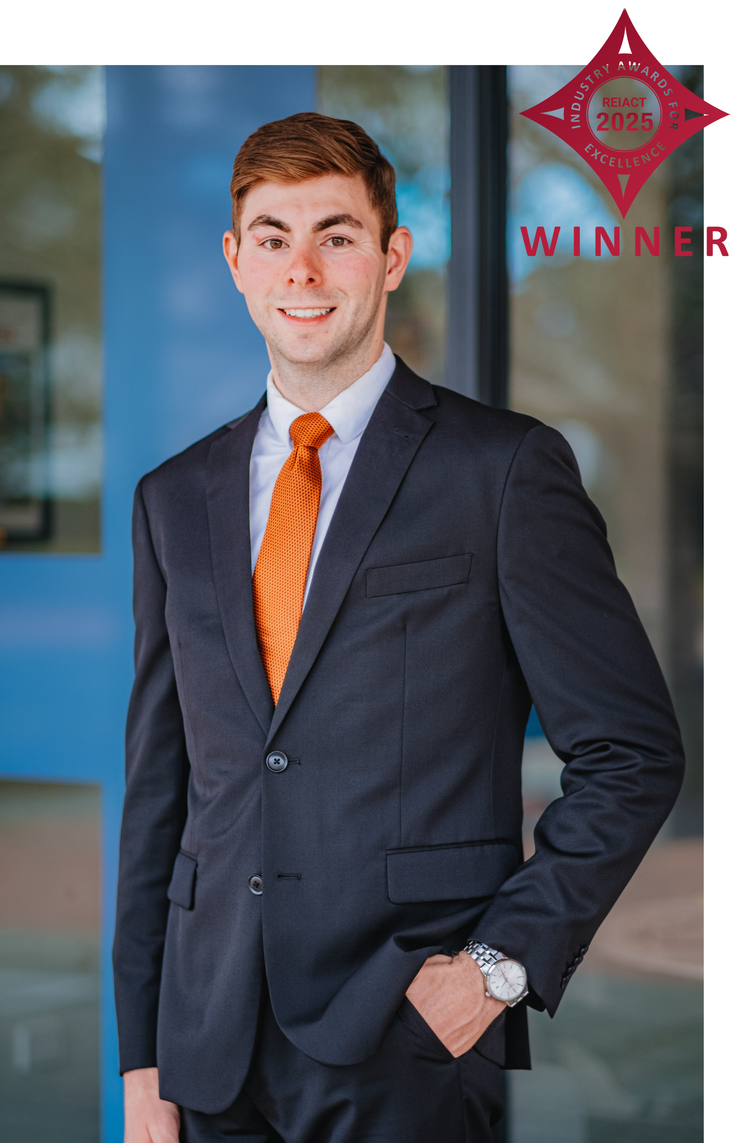 A young man with short brown hair and fair skin, dressed in a dark suit, white shirt, and orange tie, standing outdoors with a slight smile, in front of a blue wall with a window, with an award badge and text in the upper right corner.
