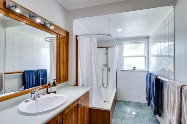 Bathroom with wooden framed mirror, white sink, and vanity, shower with white curtain, and window with potted plant, blue and gray towels hanging on towel bars.