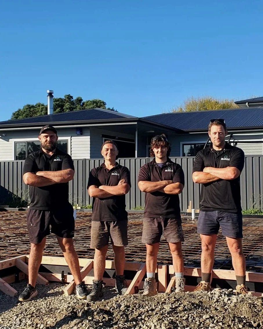 Four workers standing with arms crossed in front of a house under construction, with rebar and wooden formwork on the ground.