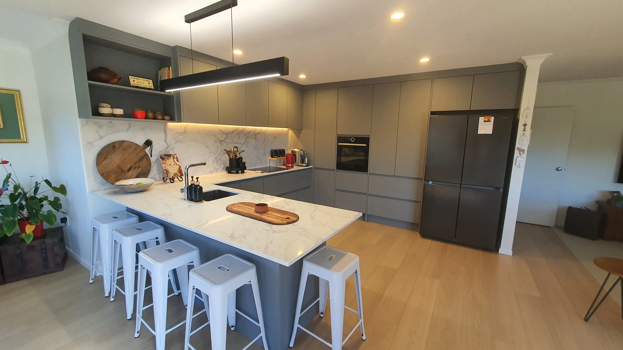 Modern kitchen with a white marble island counter, gray cabinets, built-in oven, large black refrigerator, open shelving, and bar stools.