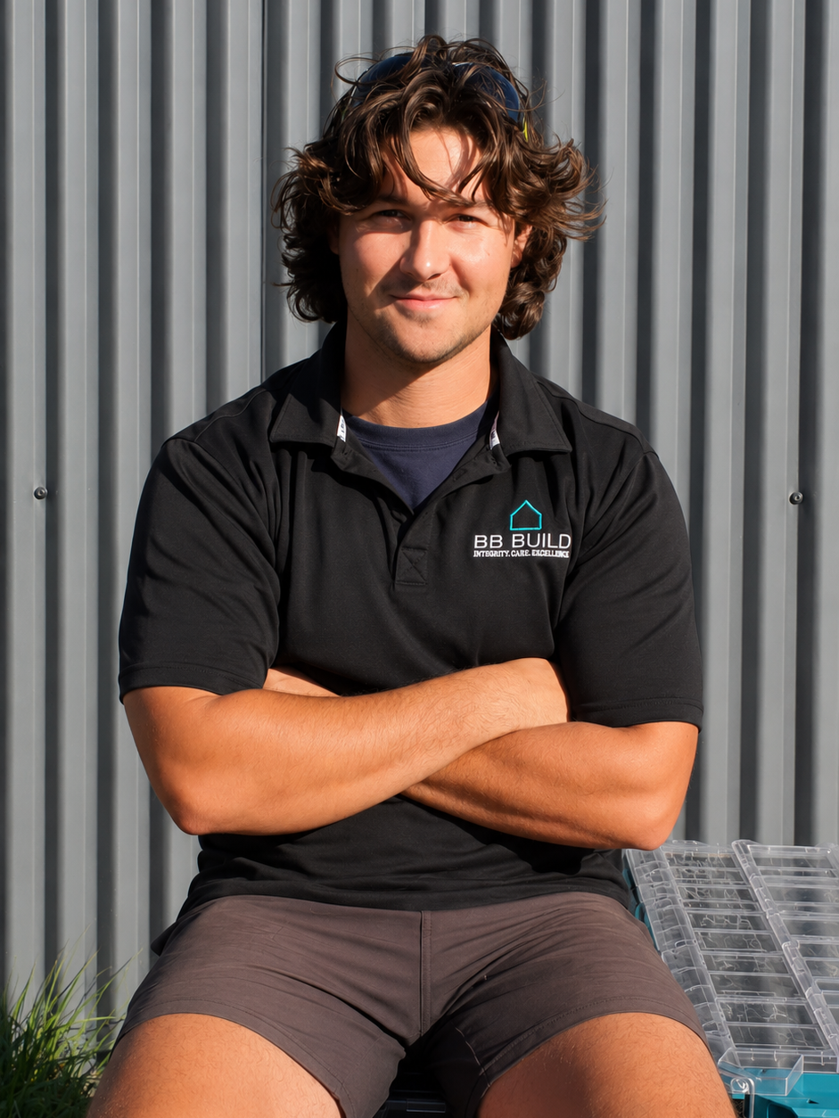 Young man with brown wavy hair sitting outdoors against a metal wall, wearing a black polo shirt with a logo and text, and shorts.