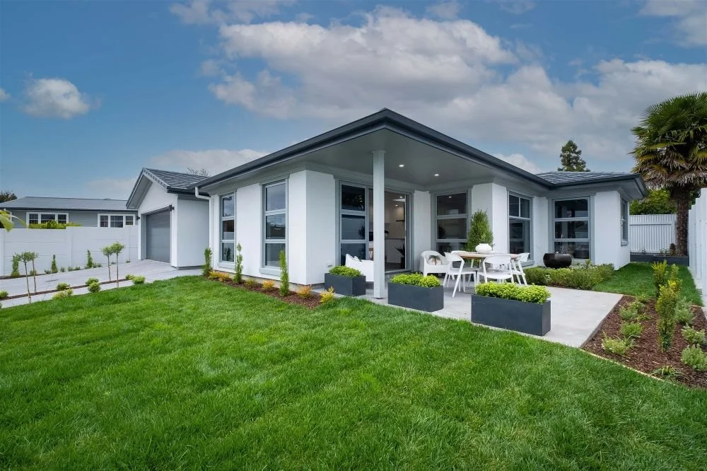 Modern white house with a large backyard, patio area with outdoor furniture, lush green grass, and a blue sky with scattered clouds.