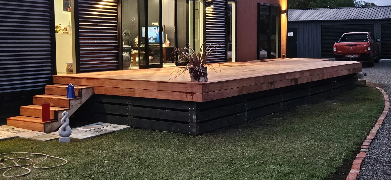 A modern wooden deck attached to a house, with stairs leading up, grass in front, and a potted plant on the deck. A black and a red car are parked nearby, and the house has large glass doors and exterior lighting.