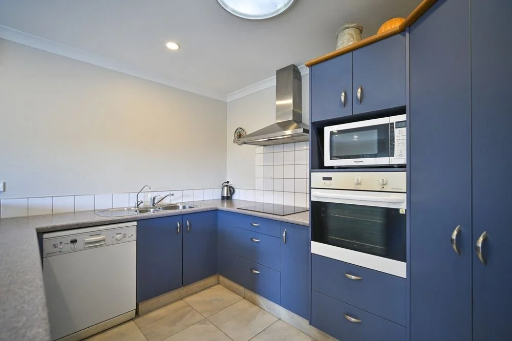 Kitchen with blue cabinets, white tiled backsplash, dishwasher, microwave, oven, stainless steel range hood, and beige countertops.