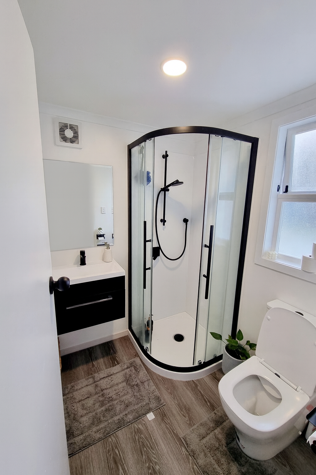 Small modern bathroom with a walk-in shower, white walls, a black vanity with a mirror, a toilet, and a window with frosted glass. There is a small potted plant and a brown rug on the hardwood floor.