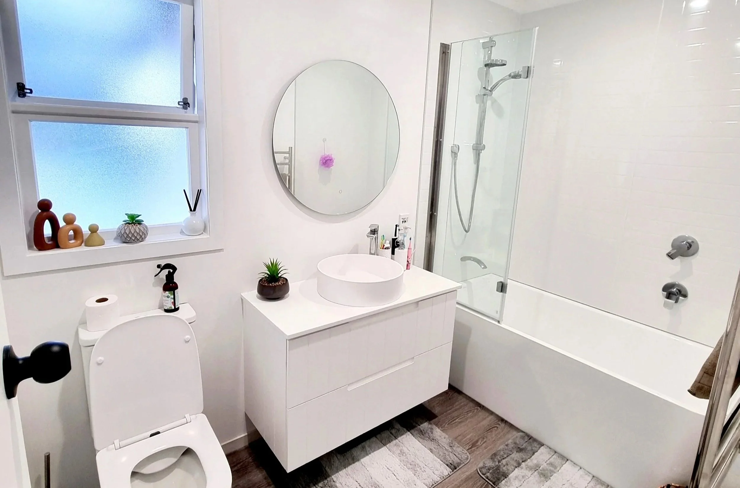A clean, modern bathroom with a white vanity, round mirror, shower with glass door, and decorative items on the windowsill.