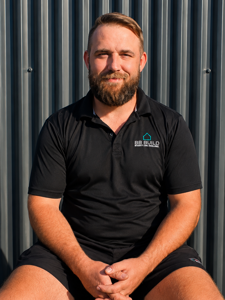 A bearded man sitting against a metal wall, wearing a black polo shirt with the logo 'BB Build' and the tagline 'Integrity, Care, Excellence.'