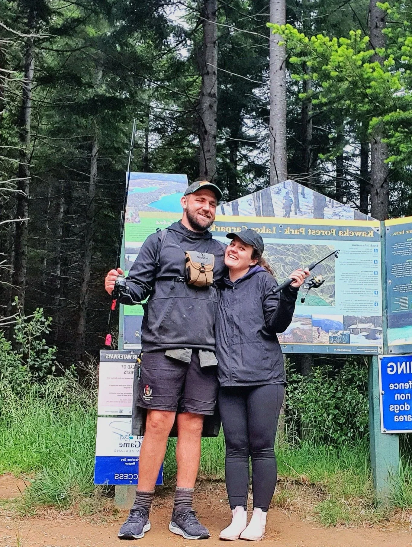 A man and a woman stand together smiling in front of an informational signboard in a wooded forest area. The man is wearing a cap, a jacket, shorts, and hiking shoes, while the woman is holding a hiking stick, wearing a cap, a jacket, leggings, and hiking shoes.