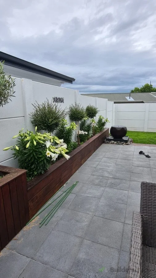 A backyard patio with gray tiled flooring, a white fence, a wooden flower bed with white lilies and shrub plants, a large decorative black pot on a stone base, and some outdoor furniture, under a cloudy sky.