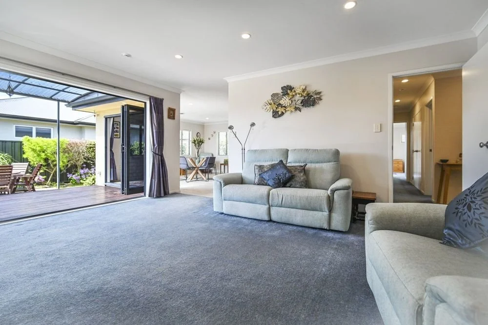 Living room with beige sofas, dark pillows, wall art, and large sliding glass door leading to a backyard patio with outdoor furniture and greenery.