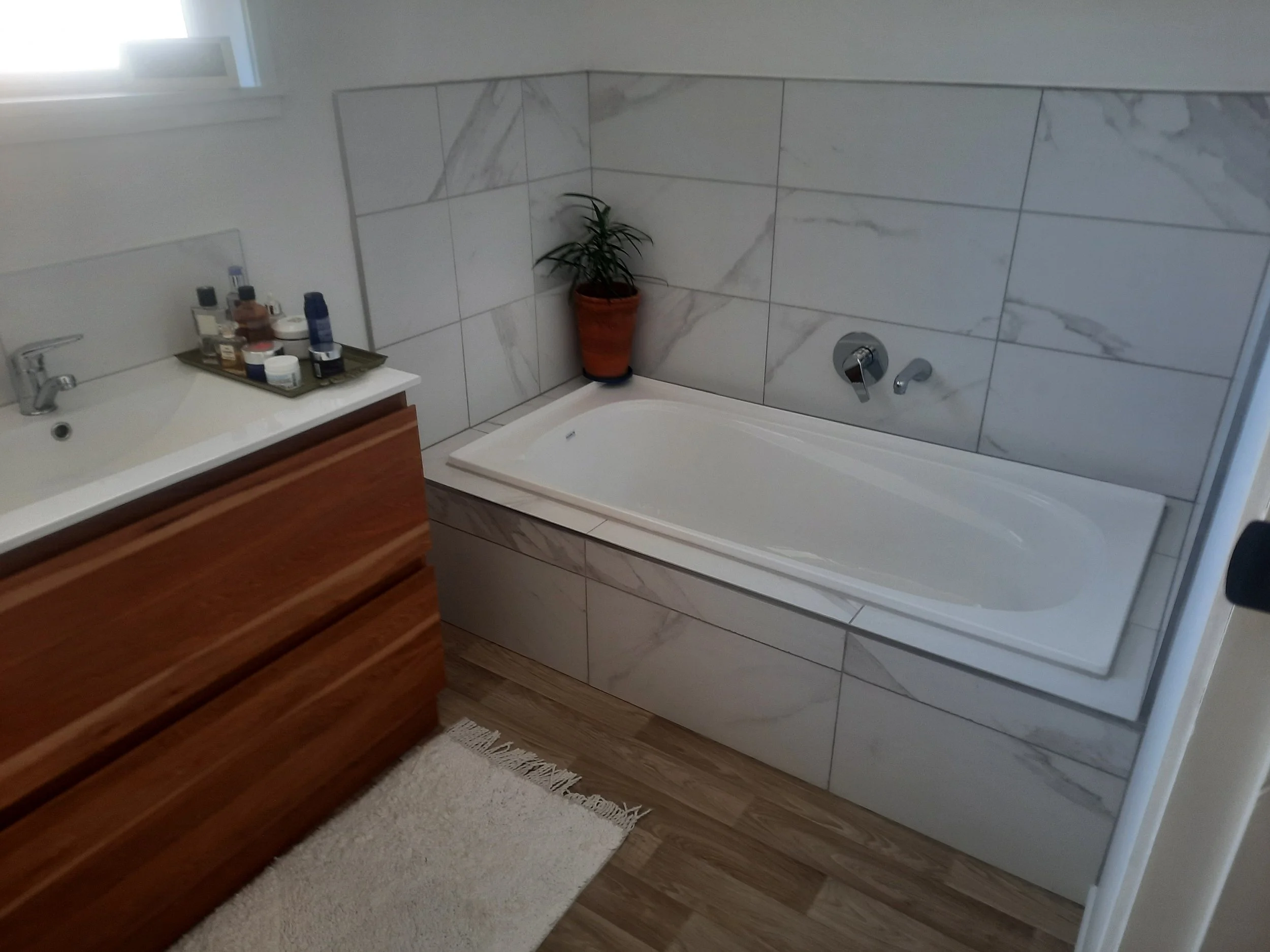 A bathroom with a corner bathtub, white marble tile walls, a potted plant next to the tub, a wooden vanity with personal care products, a beige rug, and a small window.