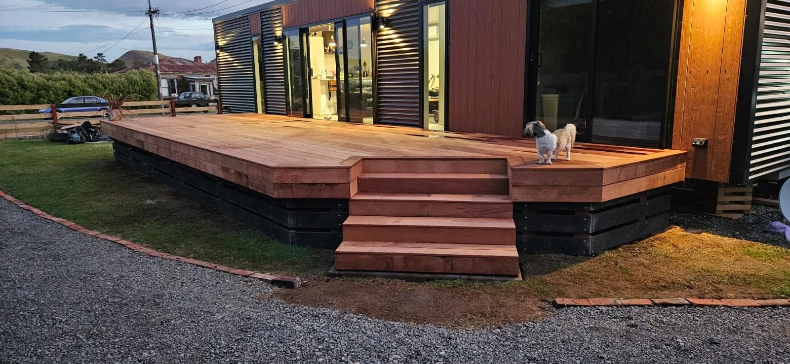 Newly built wooden deck with stairs in front of a modern house, with a small dog standing at the edge of the deck, lit by exterior lights in the evening.
